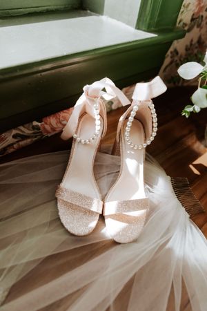 Elegant bridal heels with pearl ankle straps and glitter finish resting on tulle veil near green window. Elegant wedding in Utah.