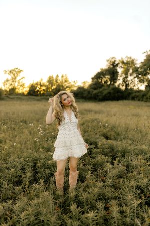 Young blonde woman in white lace dress and cowboy boots standing in golden field at sunset.