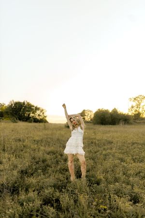 Blonde woman in white lace dress dancing joyfully in a golden sunset field with arms raised.