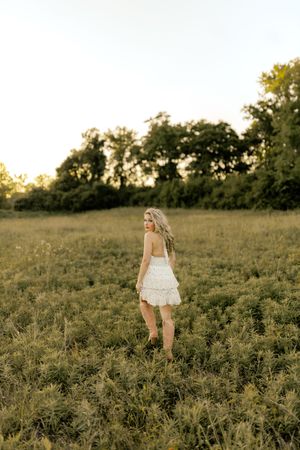 Young woman in white ruffled mini dress standing in golden sunlit meadow with lush green trees in background.