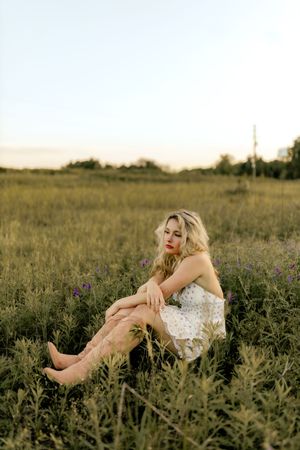 Blonde woman in white dress and cowboy boots sitting gracefully in a lush green sunset field.