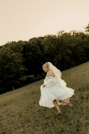 Bride in white gown and pink heels runs joyfully through a green field at sunset with trees in background. Jacksonville, NC wedding.