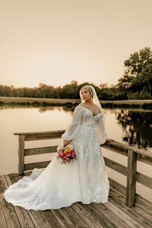 Bride in white lace gown with veil holds colorful bouquet on wooden dock at golden hour sunset.