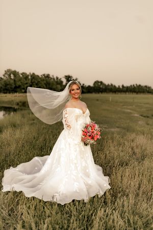 Bride in white off-shoulder gown with flowing veil holds coral bouquet in a green field at golden hour.