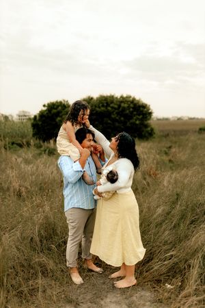 Family of four enjoying a sunny day in a grassy field, parents holding children while smiling outdoors.