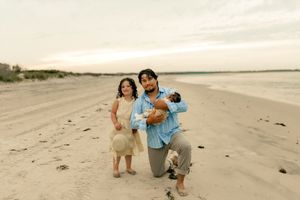 Family of three poses on sandy beach at sunset, father kneeling holding infant, mother in yellow dress beside him.