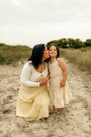 A mother kneels on sandy beach path kissing her young daughter's cheek, both wearing matching yellow dresses.