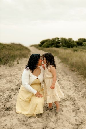 A mother and daughter in matching yellow dresses share a tender nose-to-nose moment on a sandy beach path.