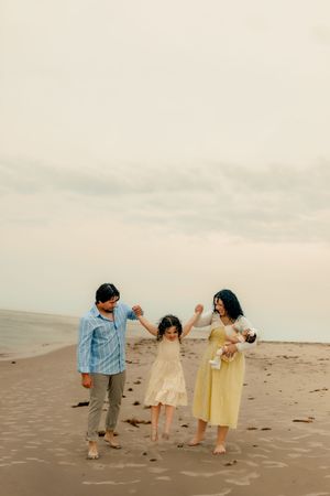 Family of four enjoying a beach day, parents swinging young daughter while mother holds baby on cloudy shoreline.