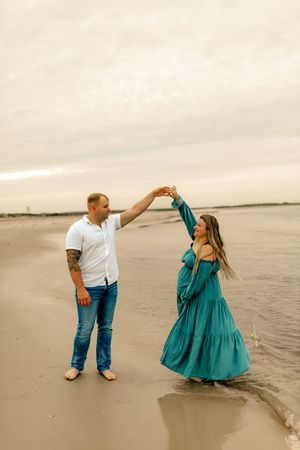 Couple dancing on beach, man in white shirt and jeans, pregnant woman in teal dress, romantic maternity photo.
