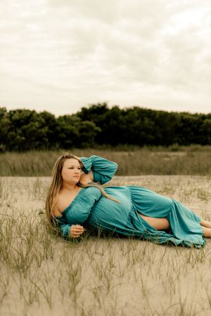 A pregnant woman in a teal dress reclines gracefully on sandy ground surrounded by wild grass and trees.