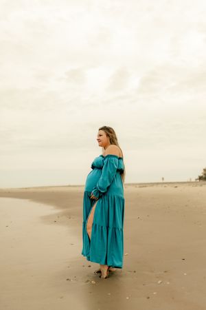 Pregnant woman in teal off-shoulder dress standing on beach, cradling baby bump, overcast sky background.