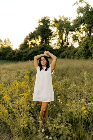 Woman in white dress standing in wildflower field at golden hour, arms raised, surrounded by yellow flowers and trees.