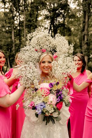 Bride in white gown holds colorful bouquet surrounded by bridesmaids in pink dresses holding baby's breath floral frames outdoors.