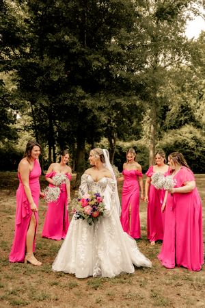 Bride in white lace gown surrounded by bridesmaids in hot pink dresses holding bouquets outdoors.