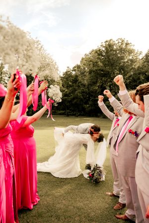 Bride and groom share a dip kiss while cheering wedding party in pink and gray attire celebrate outdoors.