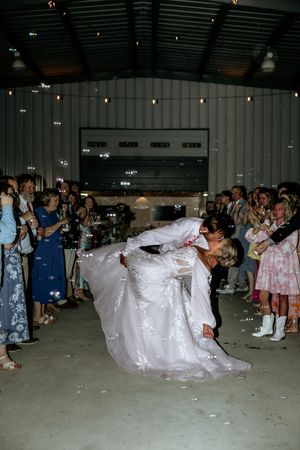 Bride and groom share a romantic dip kiss surrounded by bubbles and cheering guests at indoor wedding reception.