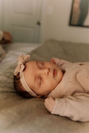 A sleeping baby wearing a beige outfit and bow headband rests peacefully on neutral bedding.