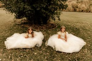 Two young flower girls in white tulle dresses sitting on green grass beneath a tree at an outdoor wedding.