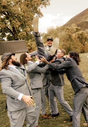 Groomsmen in gray suits lift groom outdoors with autumn mountain backdrop.