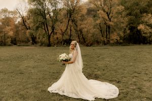 Bride in flowing white gown with veil holds bouquet in autumn meadow surrounded by colorful fall trees in Utah. Des