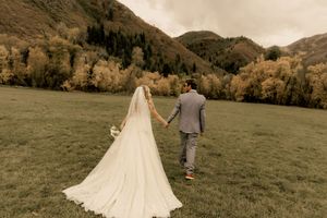 A bride and groom walk hand-in-hand across a green meadow with golden autumn mountains in the background in Utah mountains. Utah weddings.