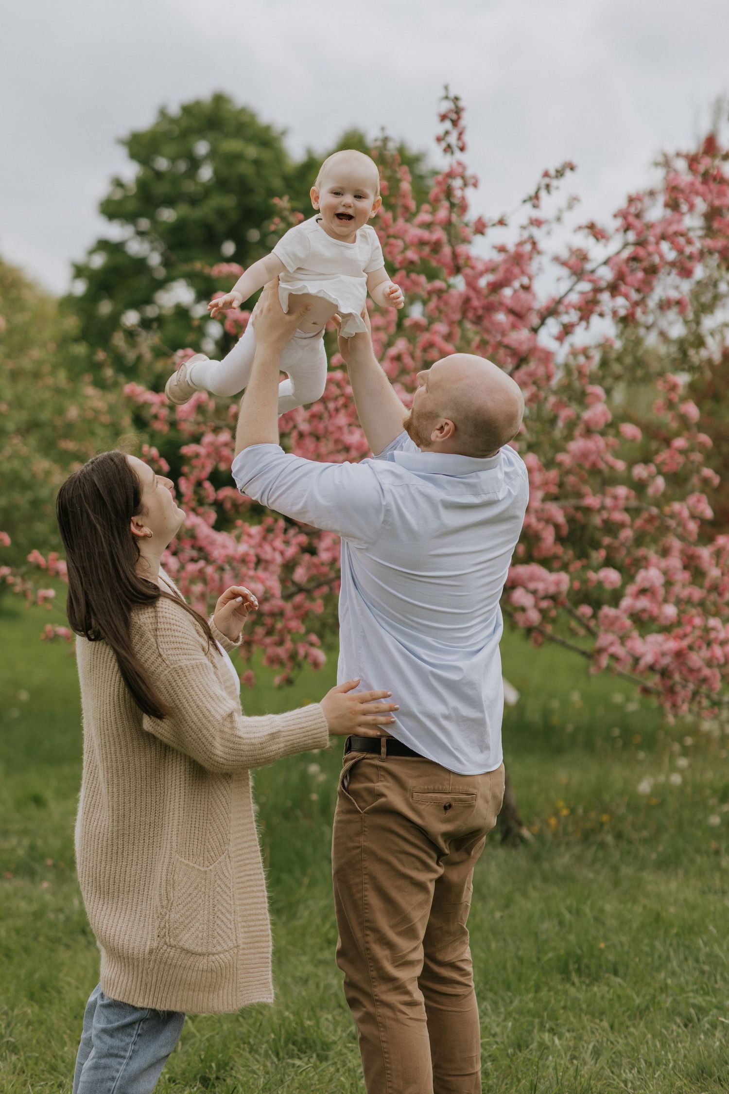 Parents playing and lifting child in the air beneath pink flowering trees.