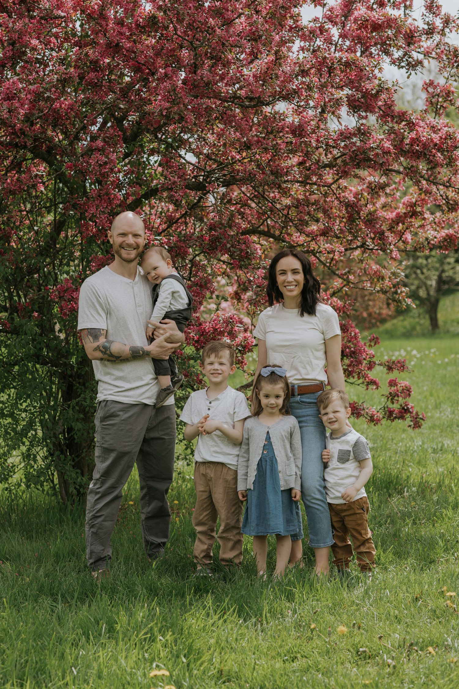 Family portrait under pink flowering trees with children in coordinated casual outfits.