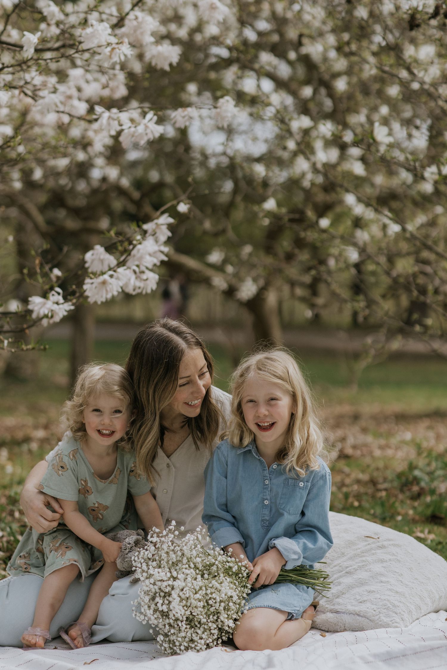 Three young friends laying on a white blanket under blooming cherry blossoms sharing joyful laughter together.
