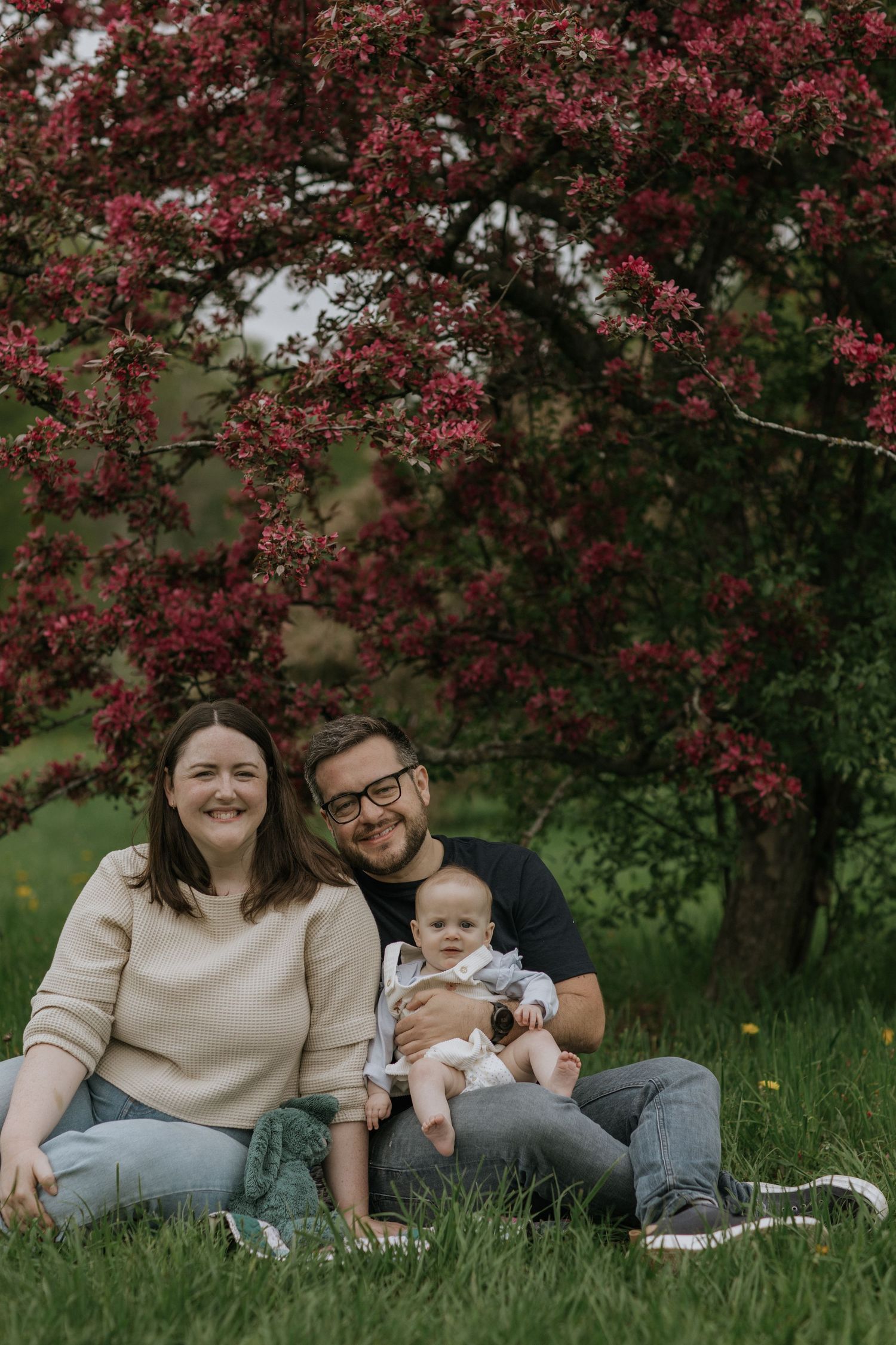 Family of three relaxing on grass under flowering tree during spring photo session.