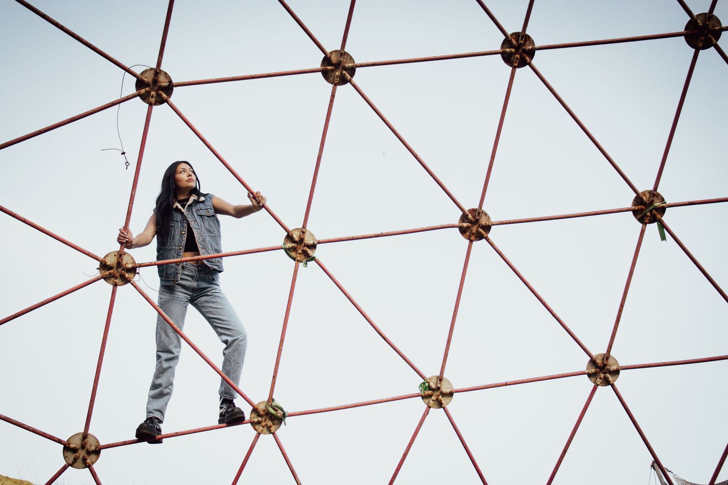 Person in casual attire climbing on geometric red metal dome structure against blue sky.