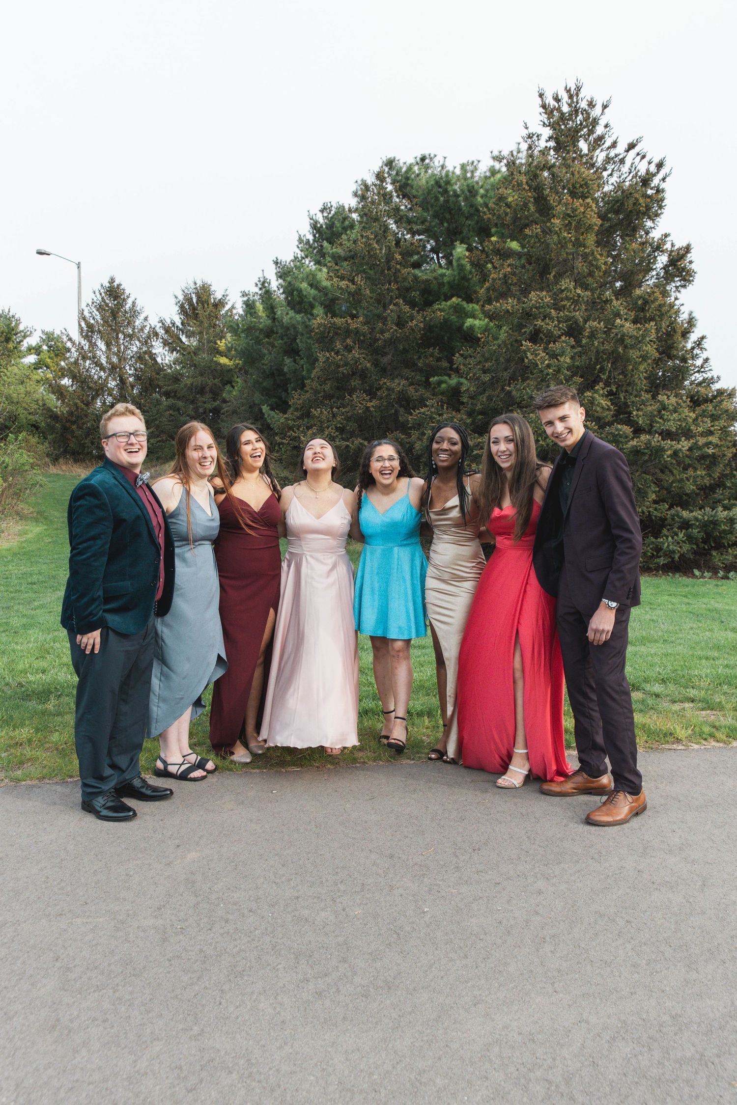 A group wearing formal attire poses together outdoors on a paved path with trees in the background.