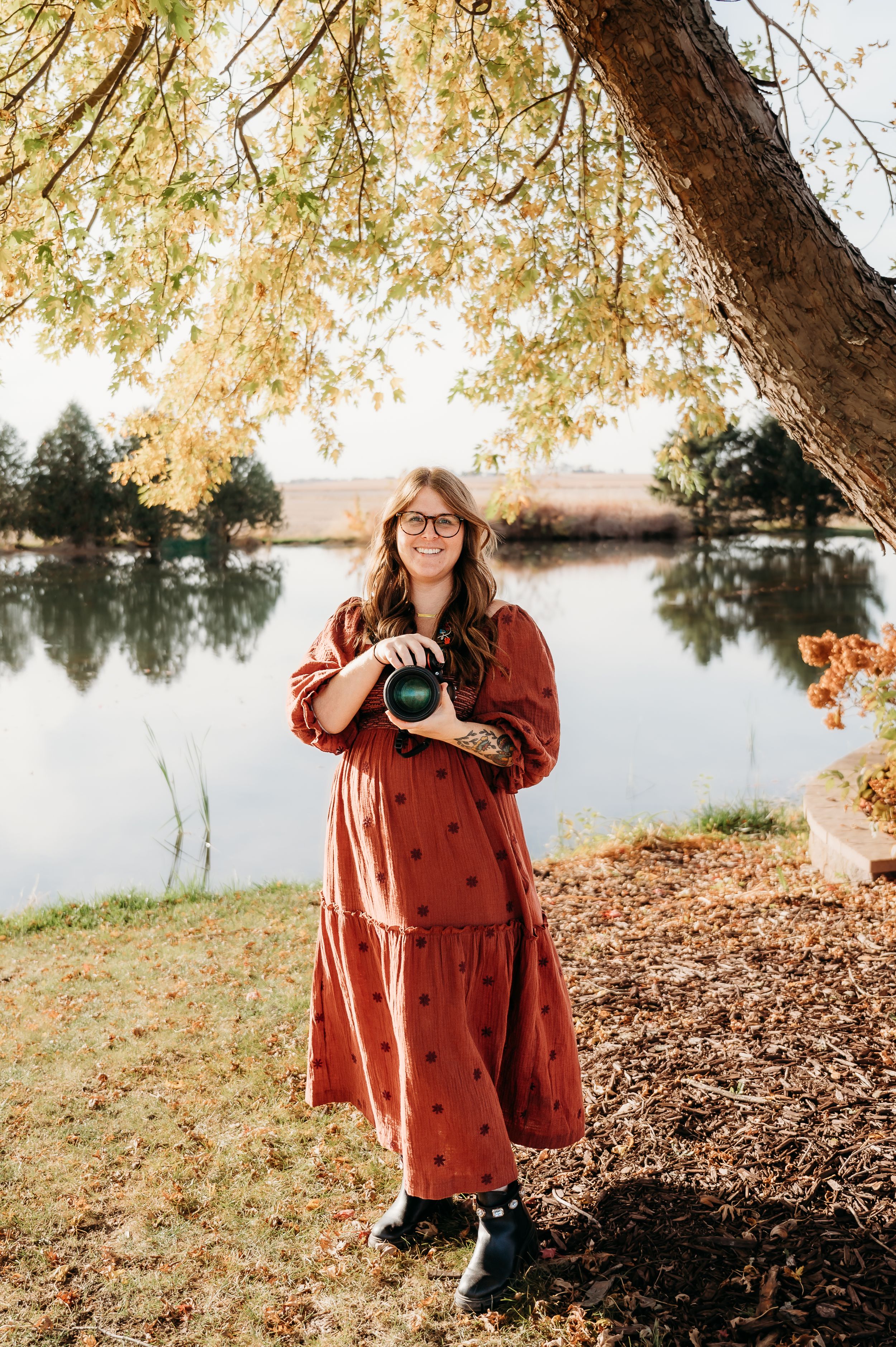 Iowa Midwest wedding and senior photographer holding camera in front of pond at Barn at Brophy Creek wedding venue. Sunny fall vibes.