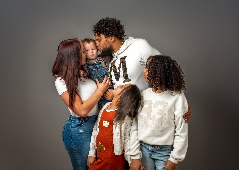 A joyful family group shares a playful moment together against a gray studio backdrop wearing casual denim and white clothing.