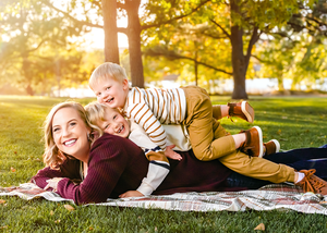 Two people lie on a picnic blanket in a sunny park with golden sunlight filtering through autumn trees.