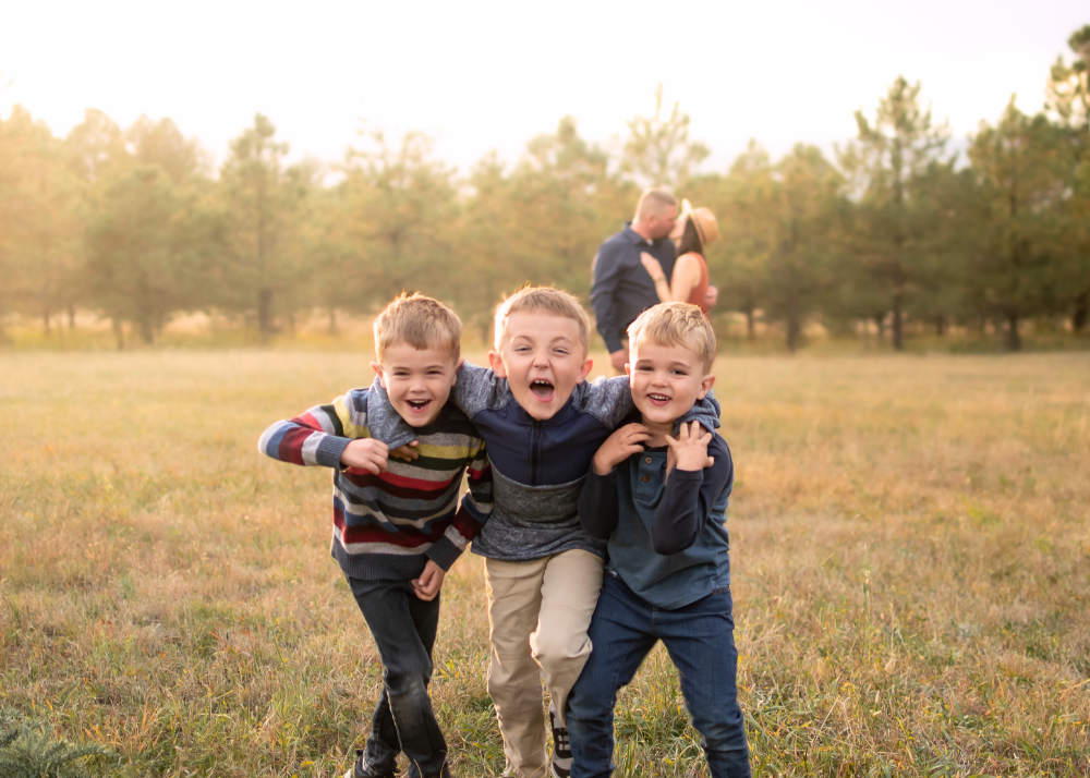 Three excited children laughing and playing together in a golden field at sunset with trees in background.