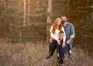 Family portrait in a rustic forest setting during sunset with tall pine trees in background.