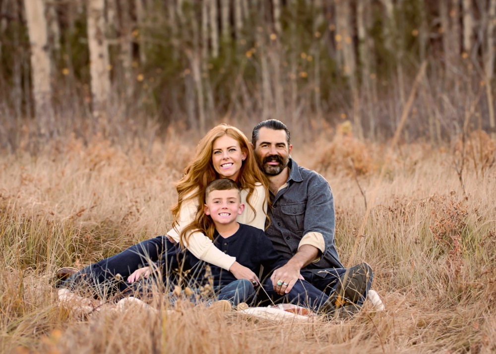 A family sits together in a golden grass field surrounded by tall trees during an outdoor photo session.