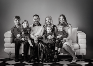 Professional black and white portrait of a family sitting together on a light-colored couch against a dark background.