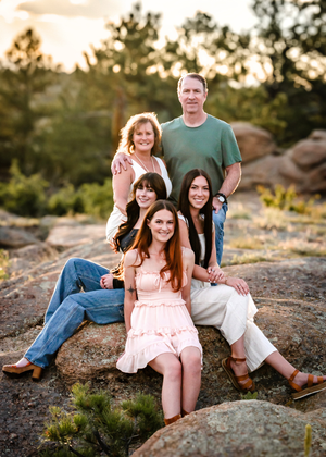 A family poses together on rocky terrain at sunset with pine trees and golden light in the background.