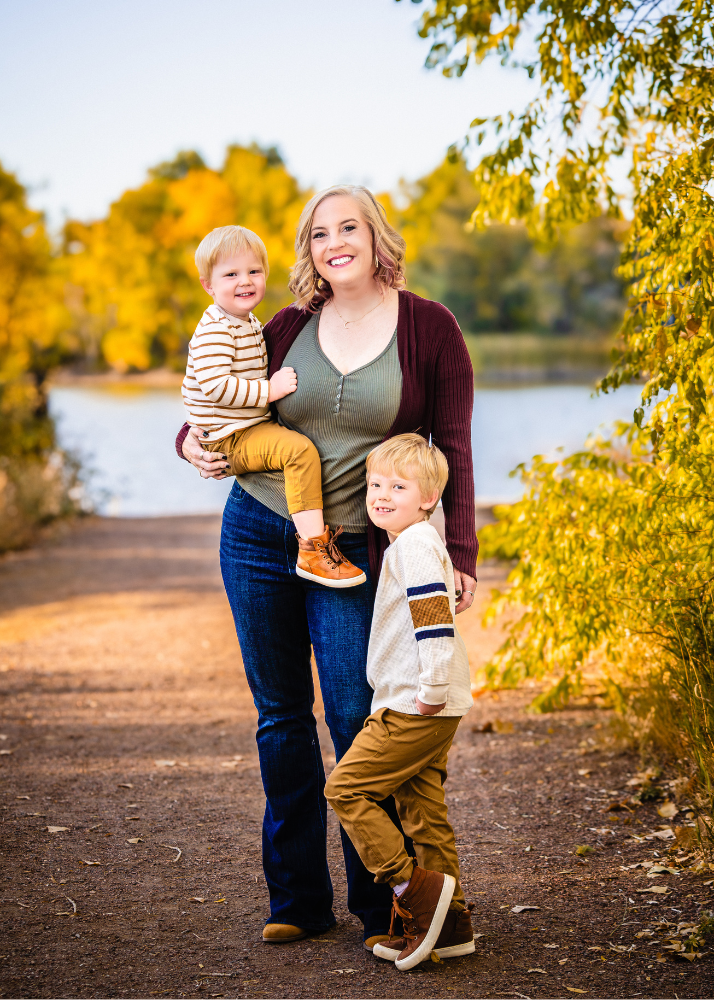 A woman and two children pose together on an autumn path by a lake with golden foliage in the background.
