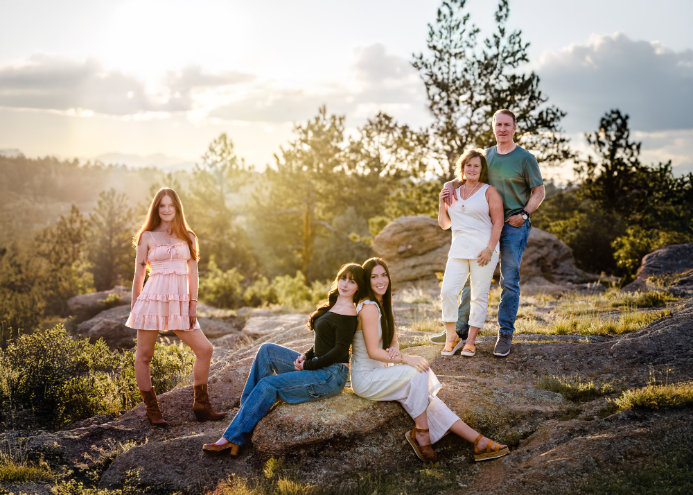 A group relaxes together at sunset on a mountain overlook with sunlight streaming through pine trees in the background.