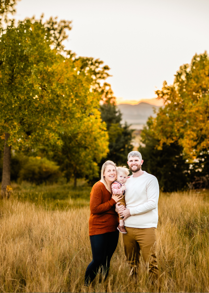 A family stands together in a golden field at sunset with autumn trees and mountains in the background.