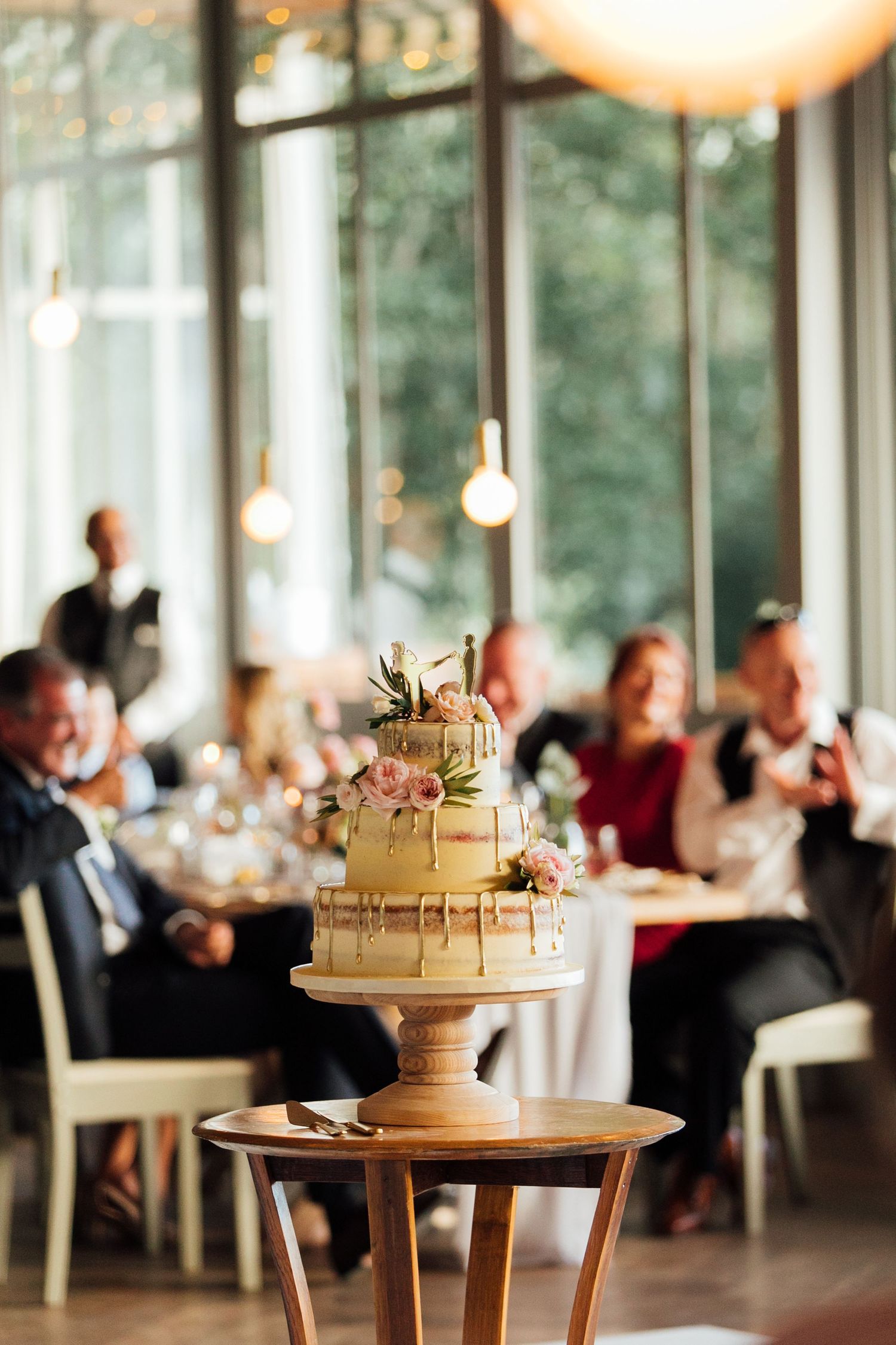 Two-tier champagne colored wedding cake with pink roses displayed on wooden pedestal table at elegant reception venue.