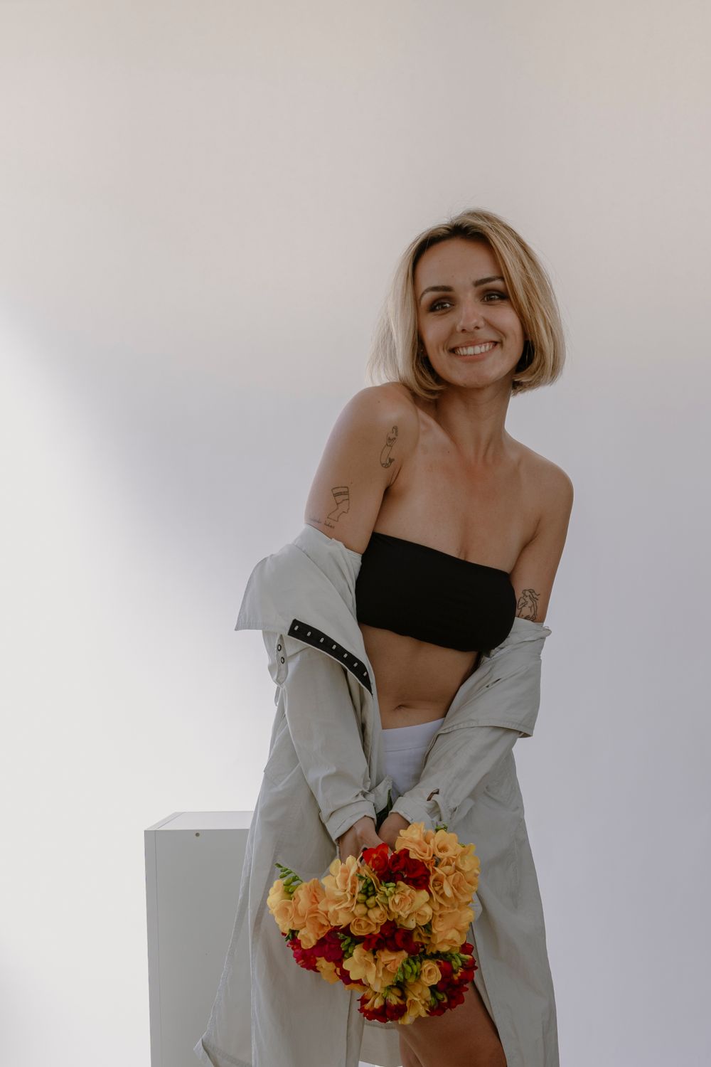 Portrait en studio sur fond blanc à la lumière naturelle. Jeune femme epaules dénudées en chemise avec un bouquet de fleurs.