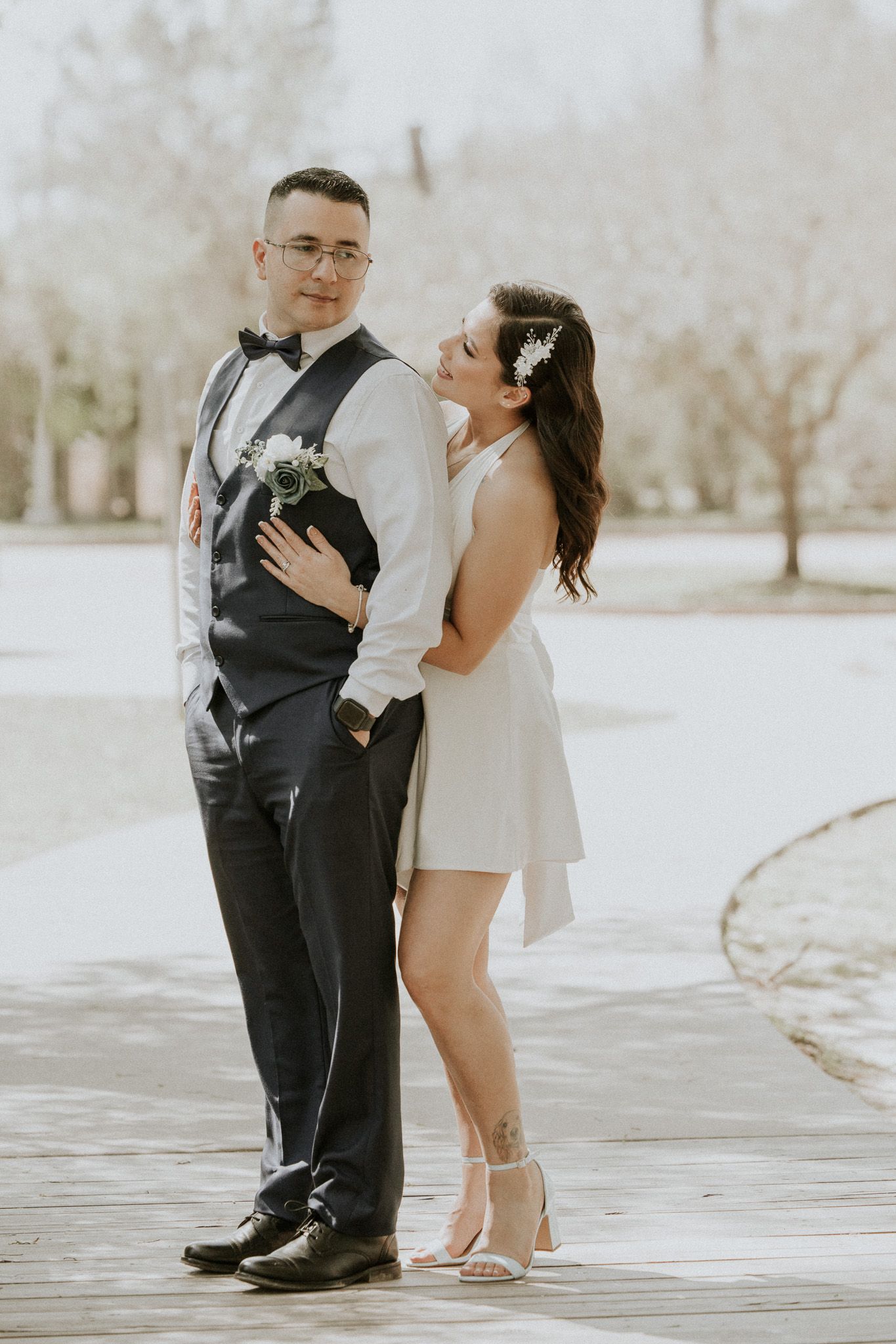 A couple poses romantically outdoors with the groom in formal attire and bride in a short white dress.