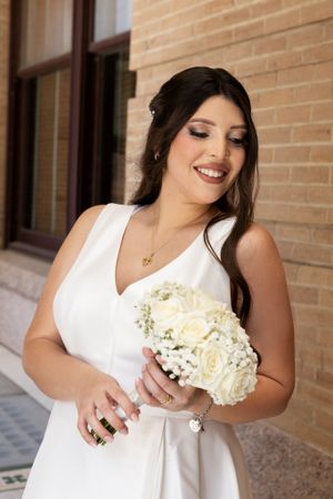 Bride in white dress holding classic white rose bouquet against brick wall.