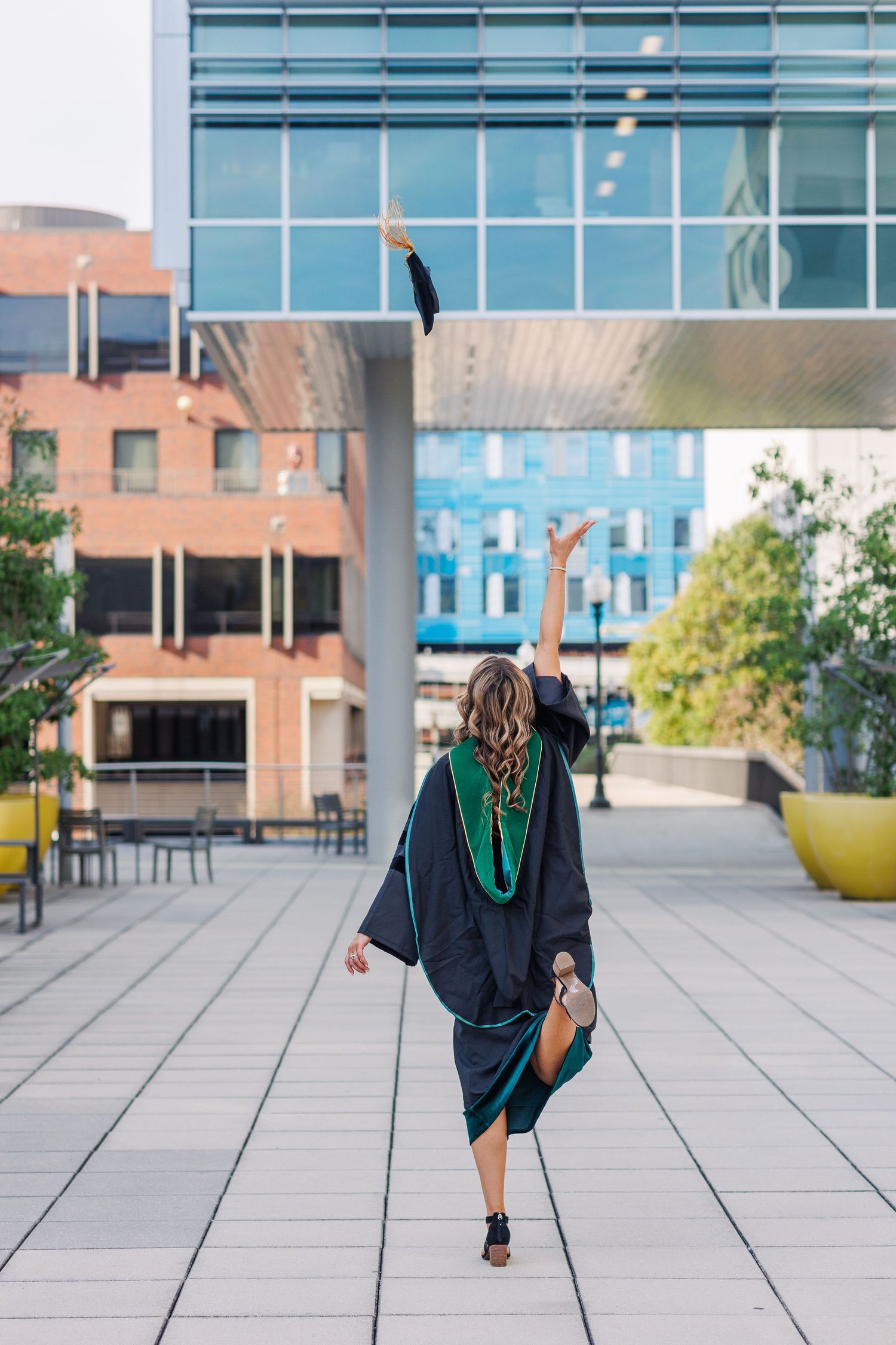 Graduate in cap and gown doing celebratory dance pose on modern campus plaza with glass building in background.