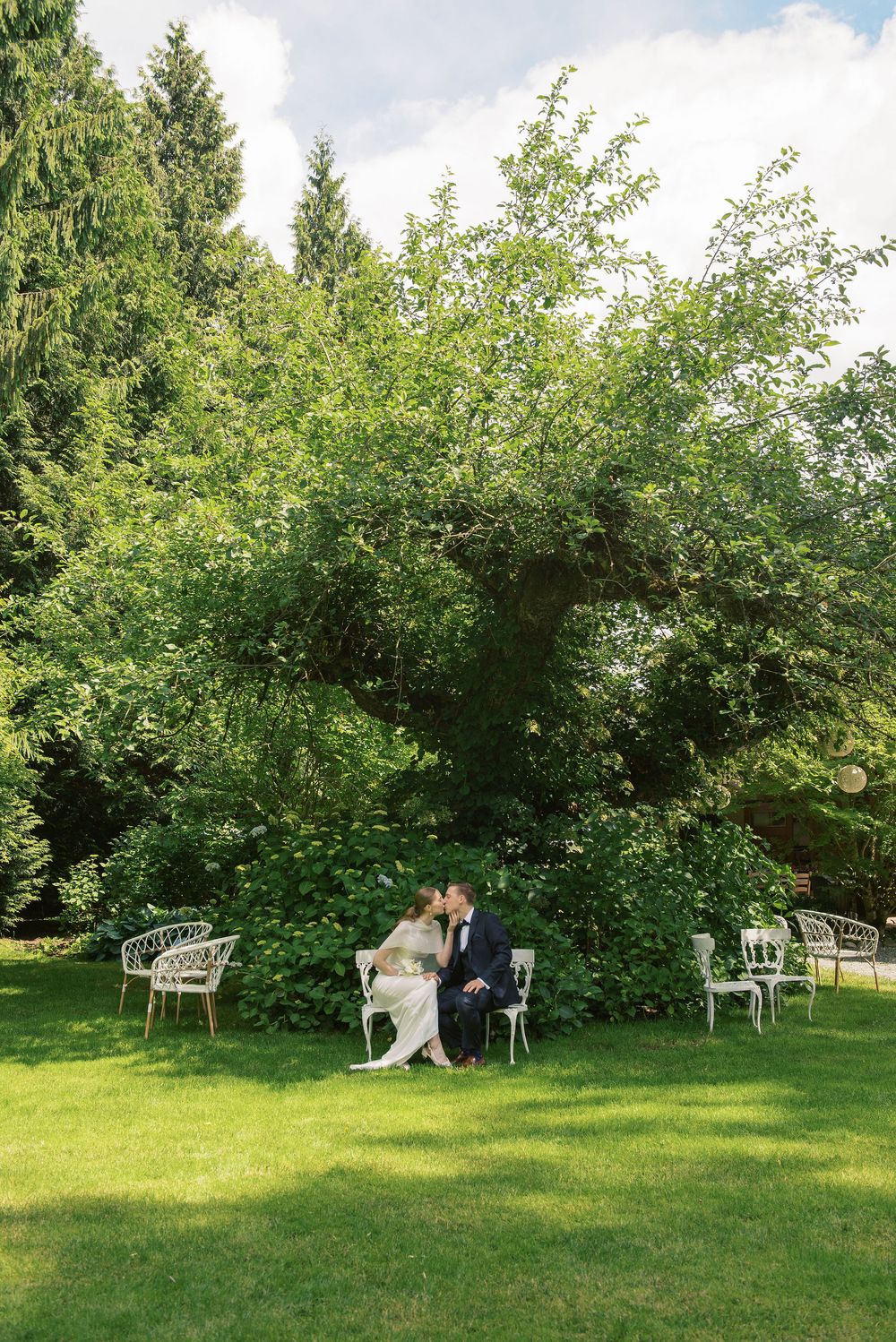 Bride and groom share a kiss on a white bench surrounded by lush green trees in an elegant garden setting.