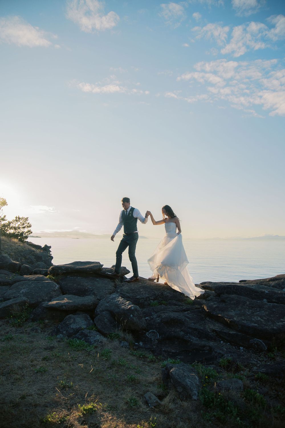 Bride and groom hold hands while walking on rocky coastal cliffs at sunset, wedding dress flowing in the breeze.